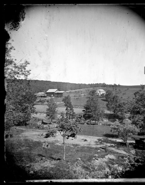 Elevated view of Andrew Amundsen's farm. A man with horses is at a well in the foreground. The well utilizes a well sweep. A barn, house and granary are on the slope of the hill beyond trees, fences and a field. Between the house and barn are a group of people in a horse-drawn wagon, and nearby two women are standing near two people on horseback.