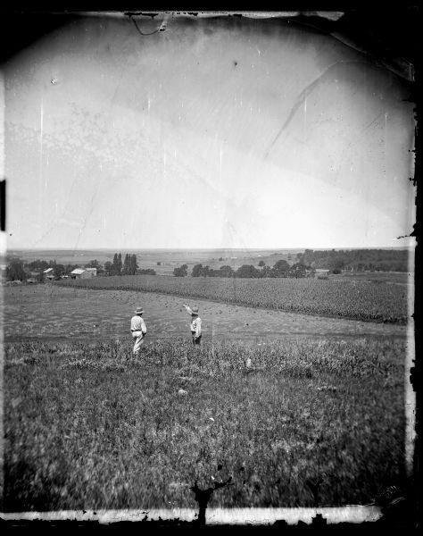 Two men wearing hats are standing in tall grass in the foreground. One man has his arm raised while pointing across a harvested field to a farmstead among trees on the left. In the distance are other farms with farm buildings and fields.