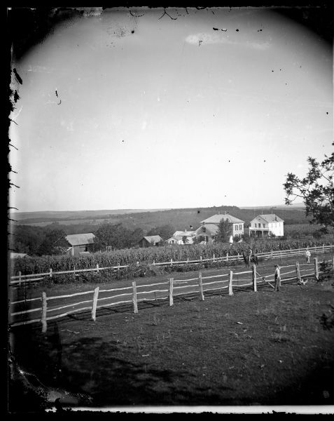 A man is standing in front of a fence along a road in the foreground. Two other men are in the road; one is standing and one is on horseback. Onon Bjorn Dahle's house and store are in the background beyond a cornfield, near the edge of a hill with farm buildings below.