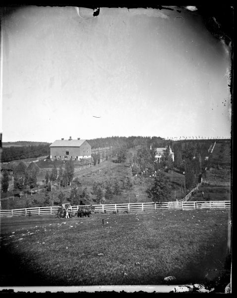 View of a prosperous farmstead from a neighboring hill. A man is standing near a woman in a horse-drawn vehicle in front of a board fence in the foreground. An elaborate system of fencing encloses haystacks; a barn with a cupola; and a barnyard with horses and pigs. A frame house, partially obscured with trees, is across from the barnyard. Men are standing with horses in front of the barn, and a man is standing with a horse behind a fence on the far right.