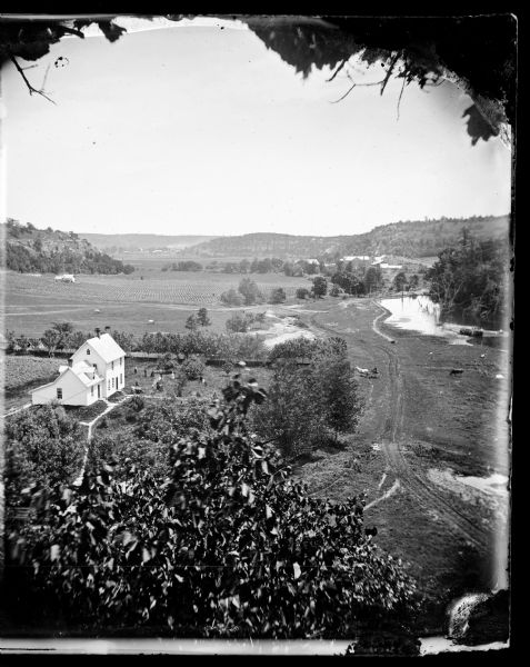 View, from above, of what may be the G. Gullockson farmstead. A frame house is in the left foreground, with a large group of men, women and children standing spaced apart in the yard. Cows are grazing in the field beyond, and other farms are in further down the valley which is surrounded by tree-covered hills. On the right a dirt road and paths lead to a pond, with more cows grazing nearby, and a man sitting in a horse-drawn carriage.