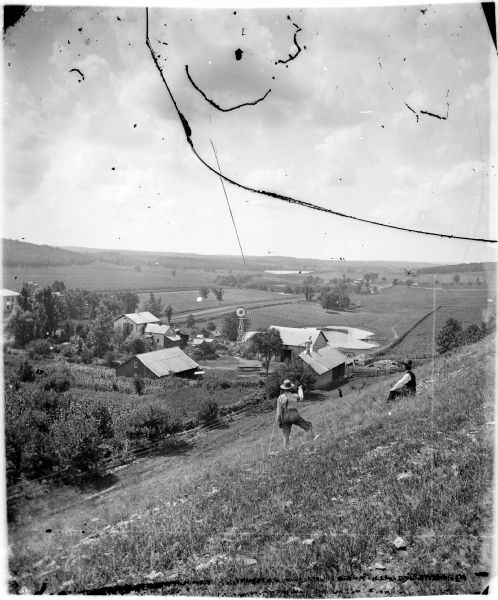 View from hill overlooking a farmstead at the base of the hill with a windmill and a pond. On the right in the foreground, two men, one sitting, one standing, have their backs to the camera and are looking out over the landscape. A fence line runs along the base of the hill, and below are the farm buildings and a farmhouse, with another farm across the road on the left. The road runs to the right towards other fields and farms. A lake is in the center in the distance.