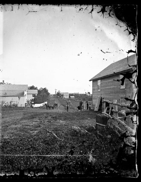 View, looking south of a man wearing a hat in a horse-drawn buggy on a rural lane in Mount Horeb. On the right a man and three children are standing near the road near a building, with a fence in the foreground. On the left two men are standing on the front landing of what may be a commercial building. Andrew Thompson's house is the second one on the left behind a fence.