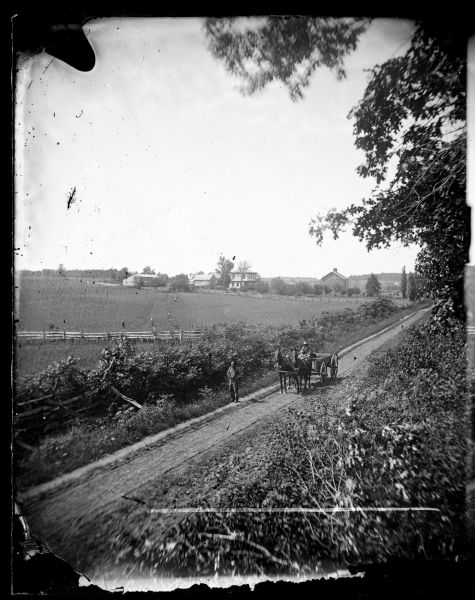 Elevated view of a man driving a horse-drawn wagon along a rural lane with a man walking in front on the left. In the background is a field, fences, farmhouse and farm buildings. People are posing spaced apart in the field behind the road. Although the location is unidentified, the fact that the photograph was taken by Andreas Dahl, a photographer known for his photographs of rural Dane County, suggests the location is somewhere in Dane County.