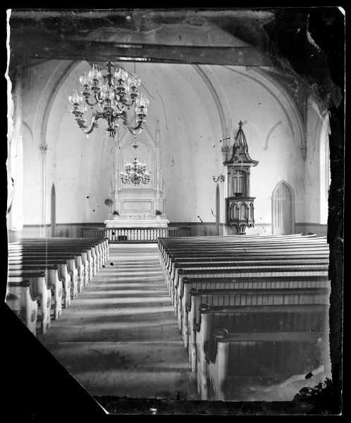 View of the First Norwegian Lutheran Church interior looking up the aisle between the pews and towards the altar. Double oil chandeliers are hanging from the ceiling, and a Norwegian communion set is on the altar.