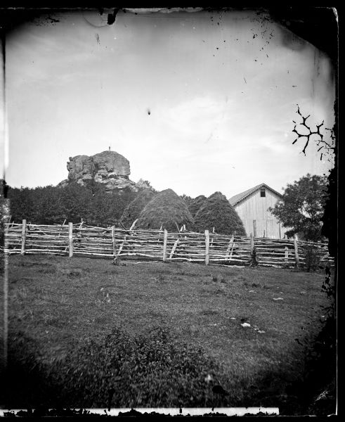 View of a farmstead with fences, haystacks and a wooden building.  A man is in the background on top of McCord Rock, also called Devil's Chimney, Preacher's Cap and Donald's Rock. I.G. Brader owned this farmstead in 1873.
