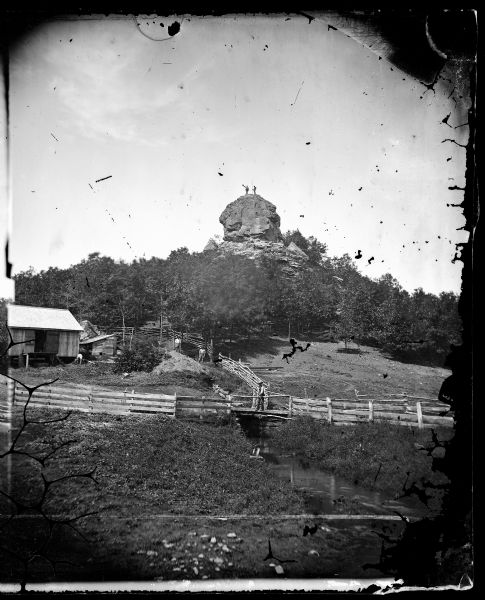 View of a farmstead with fences, haystacks and a wooden outbuilding.  Two men are posing on top of McCord Rock, one of them saluting with his hat. Another man is standing on the bridge over a river in the foreground, and two other men are standing just uphill near a tree and fencing in the center. Also known as Devil's Rock and Donald's Rock and Dahl listed the view for sale in his 1876 descriptive list as "Preacher's Cap, Springdale, Wis." The rock rises above the valley of the Sugar River.