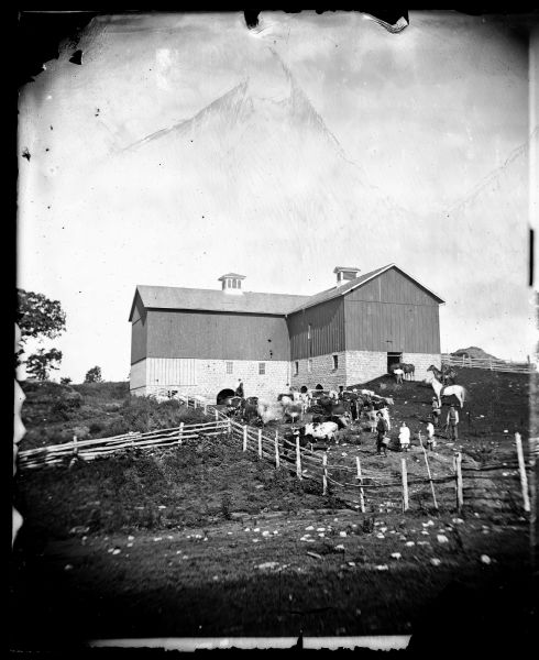 View uphill towards cattle, horses and people in the barnyard of the Tollef Gjermundsen farm. Behind them is a barn with a stone foundation with an arched entrance, and two cupolas. Men, two boys and a girl are standing in the barnyard. Three men are on horseback.