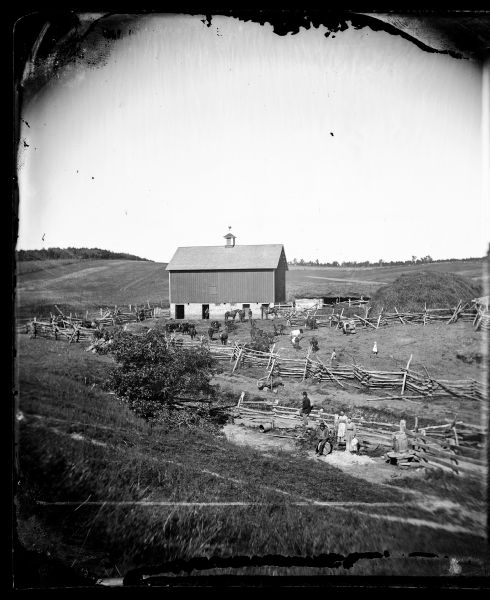 Distant view from hill of a barnyard with people, split-rail fencing, cattle, a Norwegian-style barn with cupola and rooster weather vane and an animal building with a straw roof. At the base of the hill are two women and two men posing in front of a fence. In the barnyard are children, women and men standing spaced apart among cows and horses. One man is on horseback near the barn. A field and trees are on a hill in the background.