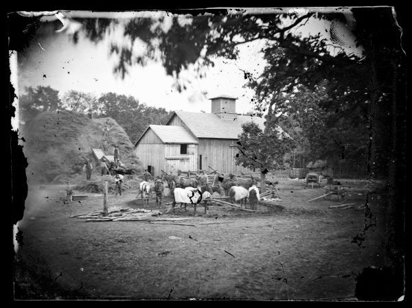 Men are in a farmyard (possibly at a sorghum mill) with horses hitched to a central-rig. A haystack, barn, wagons and farm machinery are in the background.