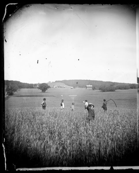 Five men, facing away from the camera, are standing in a field. Some of them are holding a rakes and a scythe. A boy is in the foreground facing the camera and shading his eyes with his hand. A farmstead is in the background. This is a carefully posed photograph.