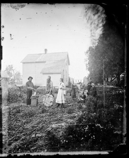 A stonecutter, a cooper or smith (identified by the tools of their trade) and a girl with a pail are standing on the left in contrast to a group of ladies and young girls sitting and standing on the right holding books and fans. There is a frame house in the background. There is a small building on the far left, and another house or large building in the far background.