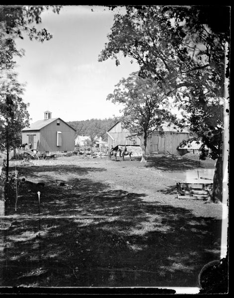 Two people are on horses in this farmyard view, including two farm buildings. One has a cupola and an unusual chute, apparently for sending hay from the loft to the feed station in the yard. The farmyard also has a hog wallow pond and an animal shade shelter. A house is in the far background.