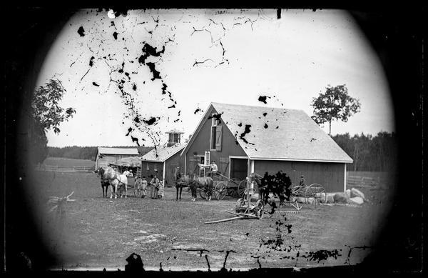 A family is in a farmyard with wagons, horses and implements. A barn with a cupola is behind them. Behind the barn is a log outbuilding with a straw roof.