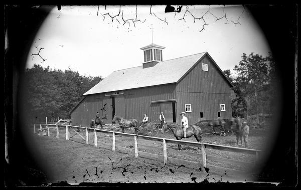 Two men are on horseback near a man sitting on a dump rake in a barnyard with the barn in the background. Another man is standing near a horse on the right, and another man is standing near one of the barn doors. Windows are cut out on the barn's lower level on the right side to shovel out the manure. There is a cupola and wind vane on the roof of the barn.