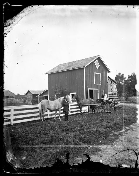 A woman and a child sitting in a horse-drawn wagon. Beside them on the left is a man standing with a horse. Behind them is a fence and a wagon shed with a diamond-shaped window near the roof, and an attached lean-to. A corn crib is in the background.