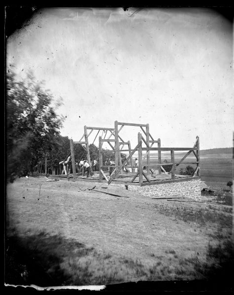 A work crew raising the bents of a barn framing system using pike poles. A valley is in the background beyond the hill where the barn is being constructed.