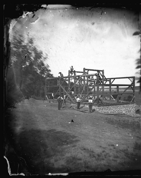 Dahl took a series of pictures of barn raisings, one of many community activities he photographed. Here the crew is installing a top plate on the side wall. A valley is in the background beyond the hill where the barn is being constructed.