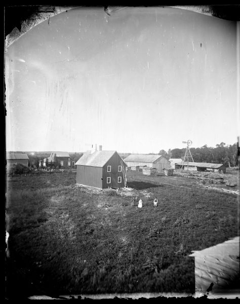 A family stands in a field with a farmstead behind them. The farmstead buildings include a hops drying shed with a large round chimney, small log structures and other miscellaneous buildings. A windmill is also in the yard.
