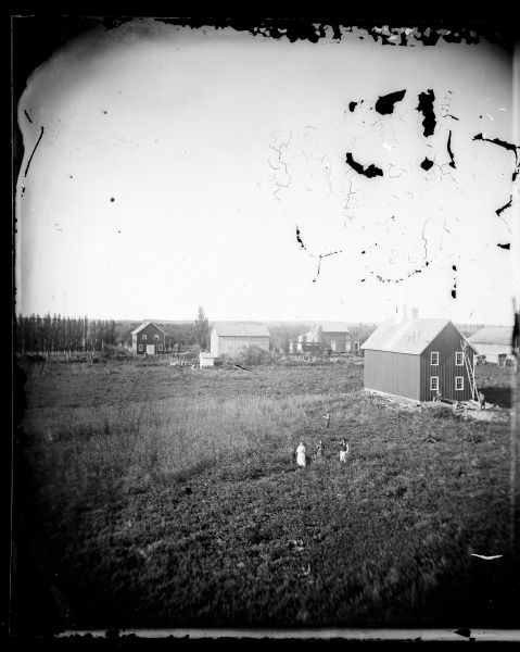 A family stands in a field with a farmstead behind them. The farmstead buildings include a hops drying shed with a large round chimney, small log structures and other miscellaneous buildings. A windmill is also in the yard.