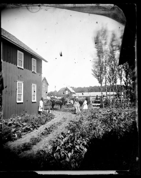 A family is at a farm in town with frame buildings, a horse-drawn buggy, and a long frame structure possibly used for drying hops.