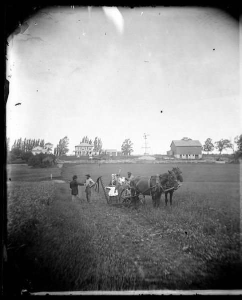 People in a field standing around a mower, with a farmstead and windmill in background, looking west. Halvor Nerison Hauge (1830-1906) purchased the farm in section 18 in 1860. It was owned in 1985 by Wayne Smithback (Breezy Heights Farm).