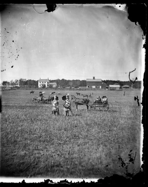 Two well-dressed women with parasols standing in a field in the foreground. Behind them are groups of people in wagons, as well as livestock, and a farmstead in the far background.