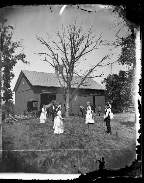 Group posed playing croquet in front of a bank barn with a handsome cupola and lightning rods. Horse-drawn wagons are in front of the barn.