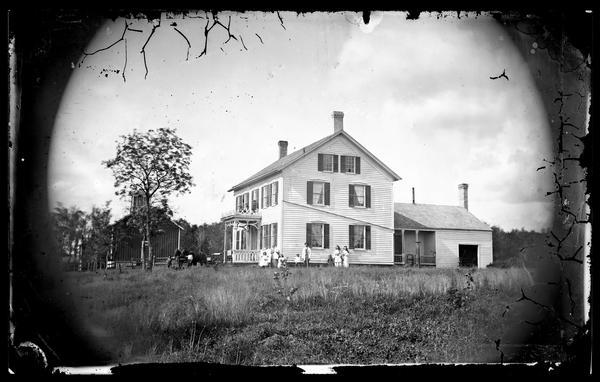 Family in yard at side of farmhouse, boy with carriage at house front, children on front porch roof. There is a barn in the background on the left, a straw topped shelter, and cattle and horses.