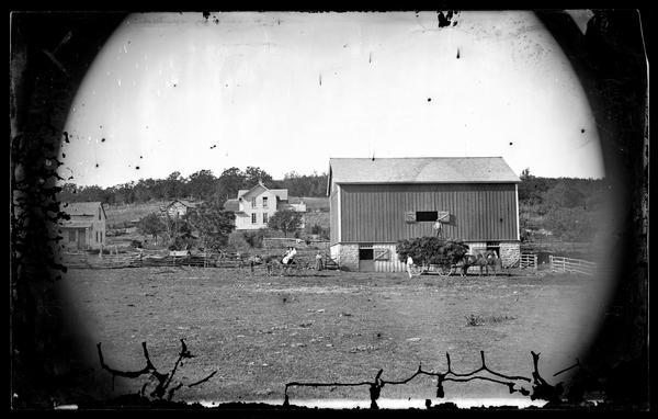 A man stands on grain bundles piled on a wagon in front of the barn, and other people are in a horse-drawn carriage on the left side of the barn. In the background are two frame houses and a log building.