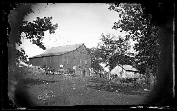 Men, horses and wagons on a hillside. A barn and another building are in the background.