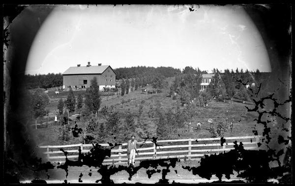 A farmer with a long beard and wearing a hat is standing in the foreground in front of a board fence, which is part of an elaborate system of fencing surrounding a farmyard. The barn is on the left, and the farmhouse is on the right. Other people are posing and standing in the background, including a group of men with horses in front of the barn, a group in the center with a horse drawn carriage, and four people in the yard below the farmhouse with croquet mallets.