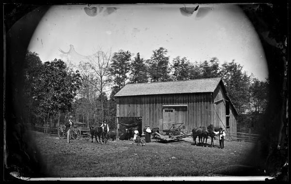A family stands in front of a barn. Beside them are cattle, horses, a wagon and a manual rake reaper.