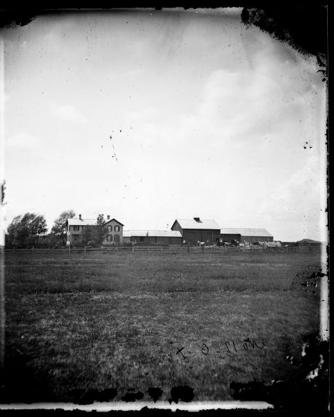 A pasture is in the foreground with a brick house, barn and connected outbuildings behind it.