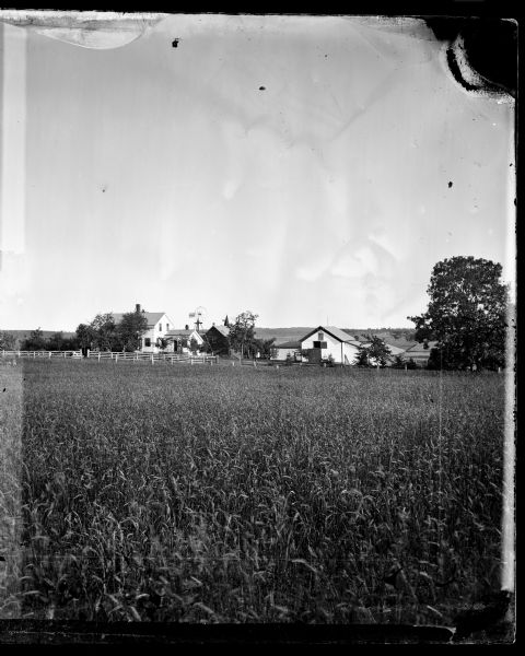 View across field with frame house, barn and windmill in background.