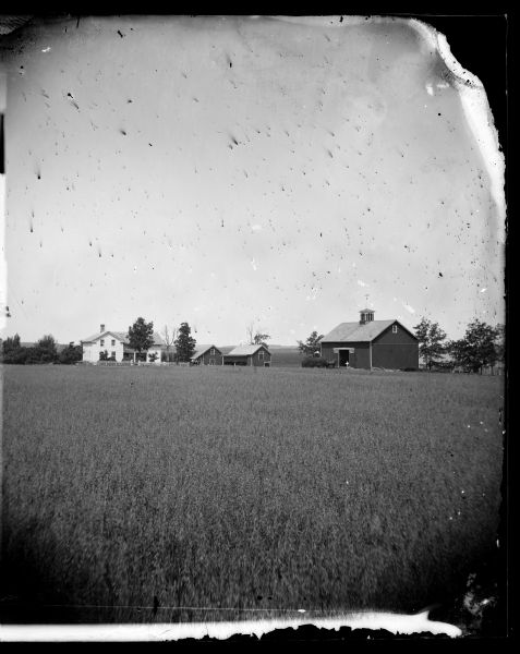 Field in foreground with house, two wood buildings and barn with man standing in front of opened doors in the background. A group of people, children?, are in a cart in the center in front of a farm building.
