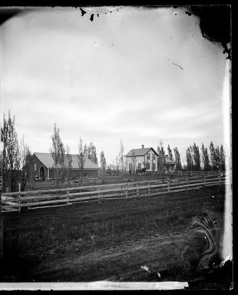 Farmyard with machinery in foreground. A man stands with a team of large horses, perhaps Percherons. Two women stand on the front porch of the farmhouse.