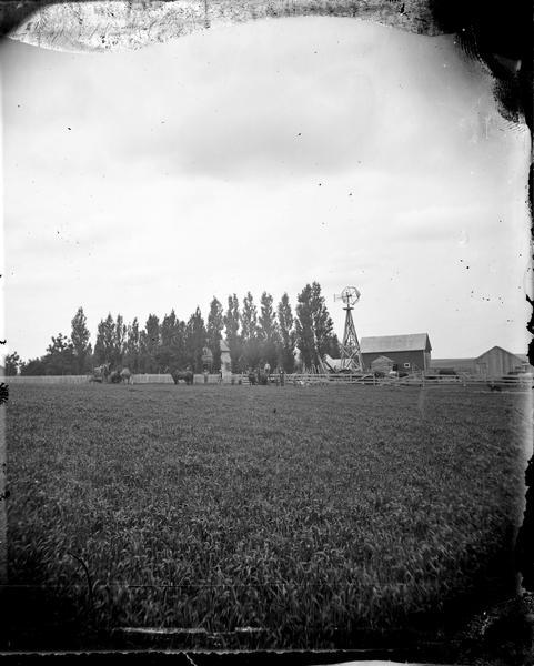 View across field, with men on farm machinery. A house, barn, windmill and woodpile are in the background.