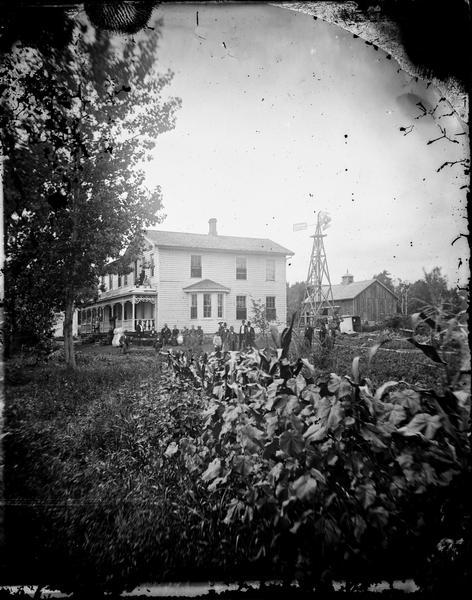 Wood frame house with carpenter's lace and family seated and standing to side of house in background. A windmill and small barn with cupola is in the right rear, and a flowerbed and stalks of corn in foreground. This is the Spring Prairie parsonage, home of Herman Amberg Preus, his wife Caroline Keyser Preus and their many children.
