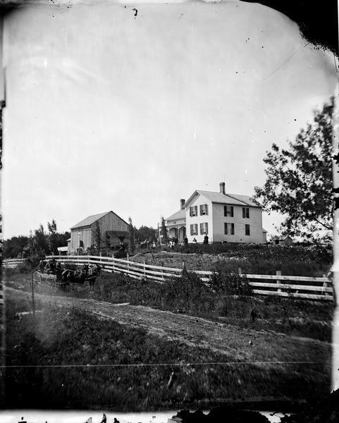 A farm adjoining a rural lane. A group of people are in a wagon at the side of the lane on the left. Beyond the fence are people on a wagon and other people standing nearby near the barn on the left, and another group of people standing in front of an upright and wing farm house on the right.