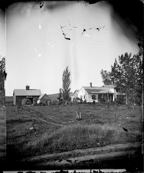 A farm house, a barn with a cupola, and a man standing on top of a haystack.