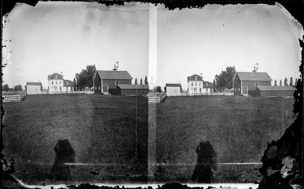 A view of the back of the Dahl residence, a second empire style house seen from across a field with a barn and other outbuildings in the background. In the foreground is the photographer's shadow.