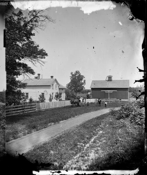 A man and two boys are standing by a road leading to a farm next to a carriage with several people sitting in it. Behind a fence on the left is a house with lightning rods and on the right is a barn with a cupola.