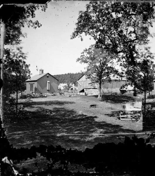 Farmyard with two barns, one with a cupola and a chute apparently for sending hay from the loft to the feed-station in the yard.  Also in this view are two horses and, in the background, a farmhouse.