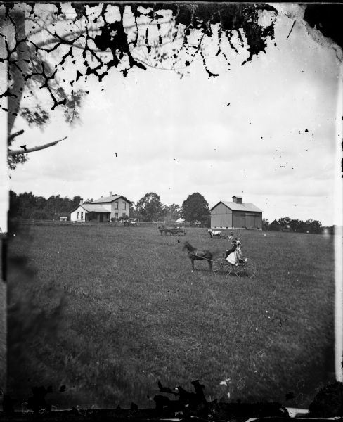 A family in a field riding on a wagon. Nearby is a hay mower drawn by a team. In the background is a granary, some haystacks behind a barn and the farmstead and brick house.