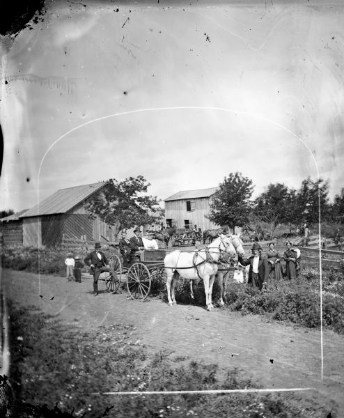 People in farmyard on horse-drawn wagon with surrounding people standing or on horseback.  Barn in background is wood frame with diagonal boards.  Note also reaper in background and drive-through corn crib.
