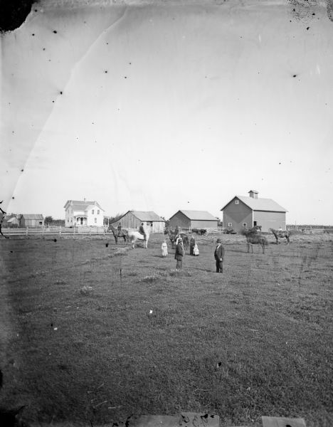 Family standing and on horseback in field with farmstead in background.