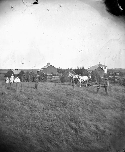 Family at work in the fields with self-raking reaper and mower, haystacks and farmstead in background.