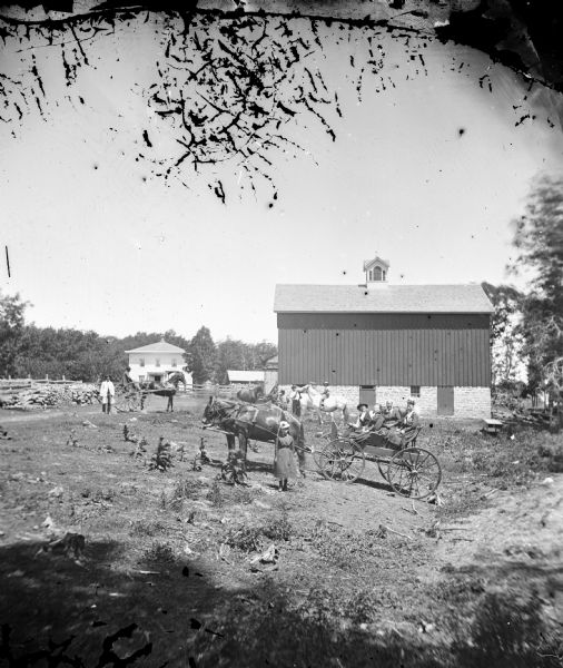 Farmyard with people in carriages, on horse, standing, with barn and house in background.  Long view of home with well.