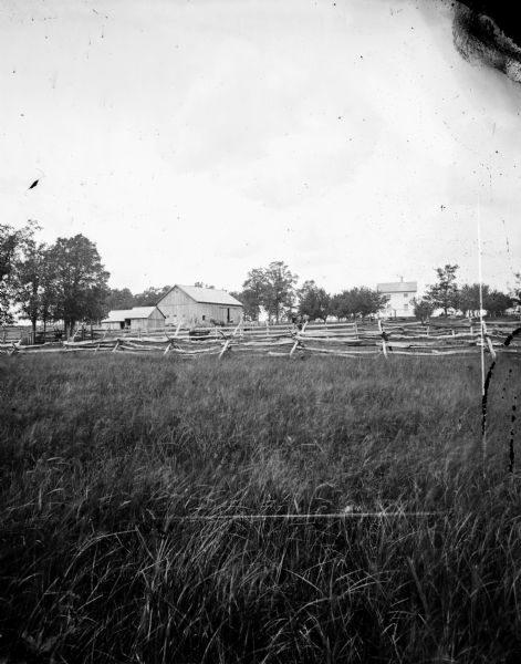 Grassy field in foreground with split-rail fence enclosing frame house, barn and small building in background. Good example of straight stake and rider fencing.
